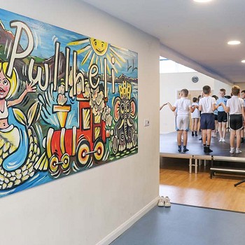 gwaith celf mawr ar y wal a myfyrwyr yn dawnsio ar y llwyfan