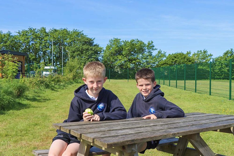 2 fyfyriwr yn eistedd ar fainc picnic, un yn bwyta afal